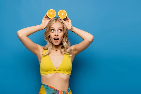 Здивована і татуйована молода жінка в жовтому бікіні зверху тримає помаранчеві половинки над головою на синьому — стокове фото