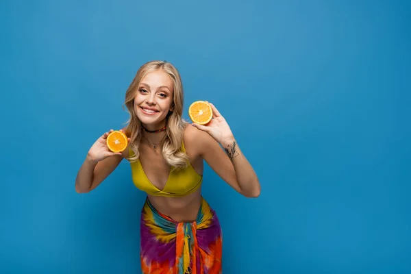 Усміхнена молода жінка в жовтому бікіні зверху тримає помаранчеві половинки на синьому — стокове фото