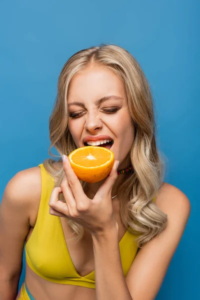 Mujer joven en bikini amarillo superior mordiendo naranja aislado en azul - foto de stock