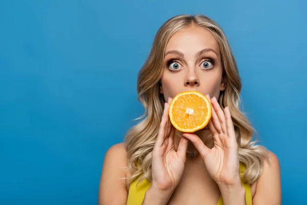 Choqué jeune femme couvrant la bouche avec orange moitié isolé sur bleu — Photo de stock