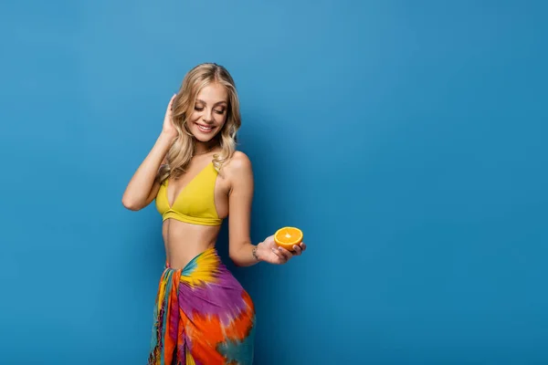 Femme blonde gaie en haut de bikini jaune et sarong tenant moitié orange sur bleu — Photo de stock