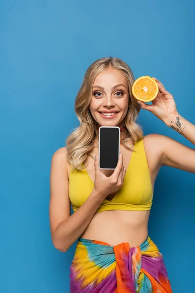 Щаслива молода жінка в жовтому верхньому бікіні тримає помаранчеву половину і смартфон з порожнім екраном ізольовано на синьому — стокове фото