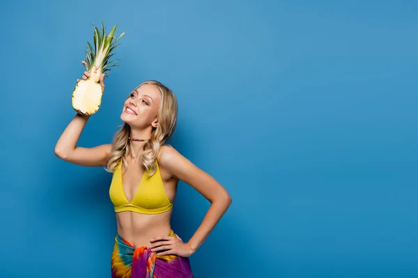 Smiling young woman in yellow bikini top and sarong holding sweet pineapple half on blue — Stock Photo