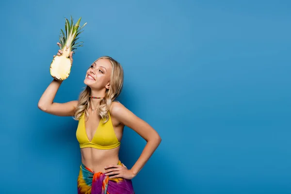 Lächelnde junge Frau in gelbem Bikini-Oberteil und Sarong mit süßer Ananas halb auf blau — Stockfoto