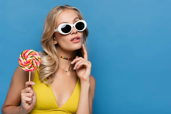 Pensive young woman in yellow bikini top and sunglasses holding sweet lollipop isolated on blue — Stock Photo