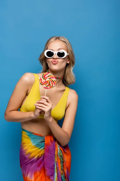 Junge Frau im gelben Bikini-Oberteil und Sonnenbrille schmollende Lippen, während sie süßen Lutscher isoliert auf blau hält — Stockfoto