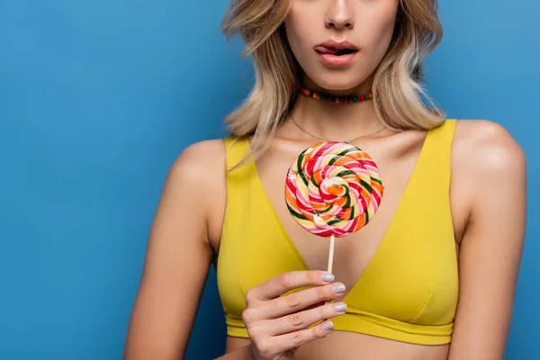 Cropped view of young woman in yellow bikini top sticking out tongue and holding sweet lollipop on blue — Stock Photo