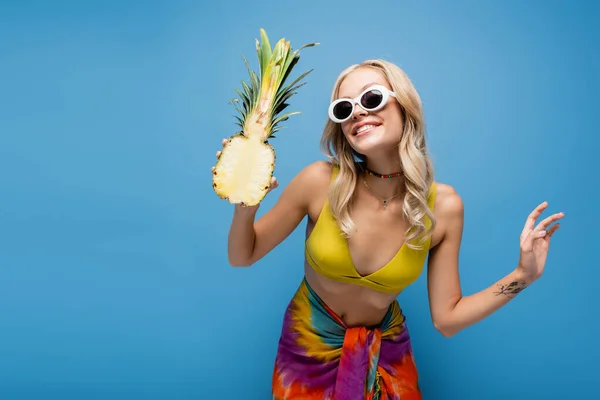 Smiling young woman in sunglasses and bikini top holding sweet pineapple half isolated on blue — Stock Photo