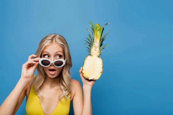 Здивована молода жінка в сонцезахисних окулярах і бікіні зверху тримає солодкий ананас наполовину ізольовані на синьому — стокове фото