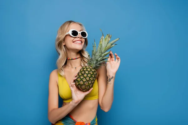 Tattooed woman in sunglasses and bikini top holding ripe pineapple while smiling isolated on blue — Stock Photo