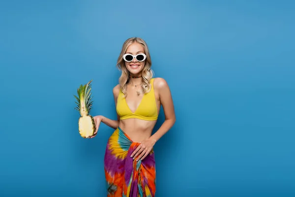 Mulher alegre em óculos de sol e biquíni top segurando abacaxi doce metade isolado em azul — Fotografia de Stock