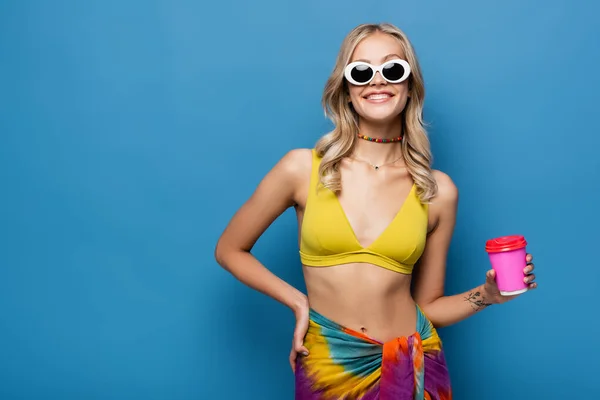 Joyeuse jeune femme en lunettes de soleil et haut de bikini tenant tasse en papier sur bleu — Photo de stock