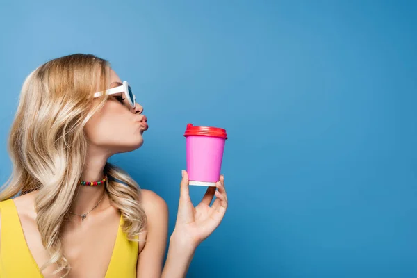 Vista lateral de mujer joven en gafas de sol y bikini superior pucheros labios mientras sostiene taza de papel aislado en azul - foto de stock