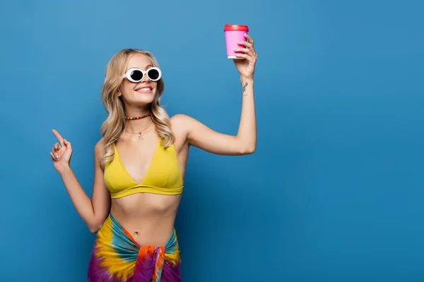Zufriedene junge Frau in Sonnenbrille und Bikini-Top mit Pappbecher auf blau — Stockfoto