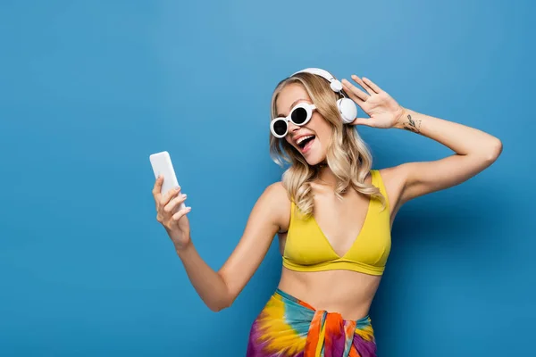 Happy woman in wireless headphones and bikini top holding smartphone on blue — Stock Photo