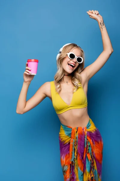 Amazed young woman in wireless headphones and bikini top holding paper cup on blue — Stock Photo