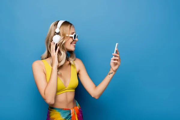 Vista laterale di donna felice in cuffie wireless e top bikini utilizzando smartphone su blu — Foto stock
