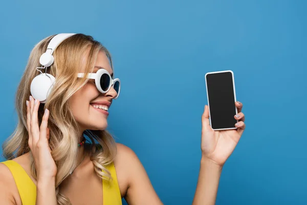Freudige Frau mit drahtlosem Kopfhörer, Smartphone mit leerem Bildschirm, isoliert auf blau — Stockfoto