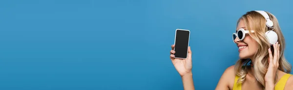 Femme joyeuse dans les écouteurs sans fil tenant smartphone avec écran vide isolé sur bleu, bannière — Photo de stock