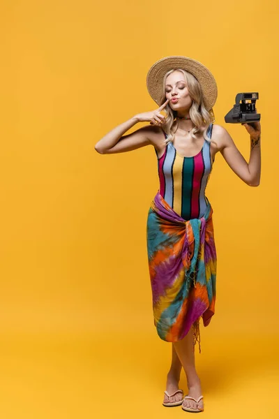 Full length of blonde woman in straw hat pouting lips and holding vintage camera on yellow — Stock Photo