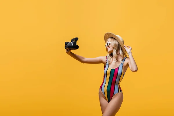 Mujer alegre en sombrero de paja y traje de baño hablando selfie en la cámara vintage aislado en amarillo - foto de stock