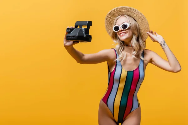 Mujer alegre en sombrero de paja y traje de baño hablando selfie en la cámara vintage aislado en amarillo - foto de stock