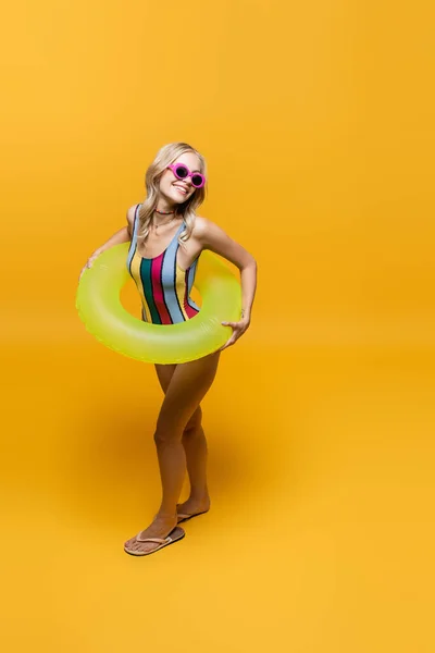 Pleine longueur de femme souriante en lunettes de soleil et maillot de bain debout avec anneau gonflable sur jaune — Photo de stock