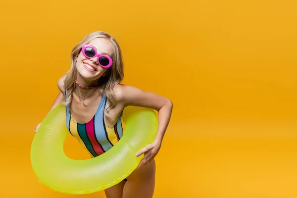 Femme souriante en lunettes de soleil et maillot de bain debout avec anneau gonflable isolé sur jaune — Photo de stock