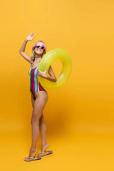 Full length of smiling woman in sunglasses and swimsuit holding inflatable ring and waving hand on yellow — Stock Photo