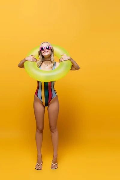 Longitud completa de mujer sonriente joven en gafas de sol y traje de baño que sostiene el anillo inflable en amarillo - foto de stock