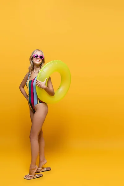 Pleine longueur de femme souriante en lunettes de soleil et maillot de bain tenant anneau gonflable et posant sur jaune — Photo de stock