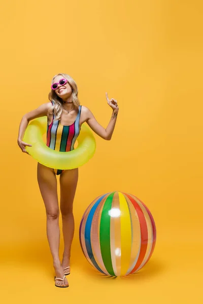 In voller Länge lächelnde Frau in Sonnenbrille und Badeanzug, die mit aufblasbarem Ring in der Nähe des Strandballs steht und auf gelb zeigt — Stockfoto