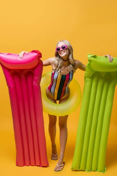 Full length of cheerful woman in sunglasses and swimsuit holding inflatable mattresses and standing with swim ring on yellow — Stock Photo