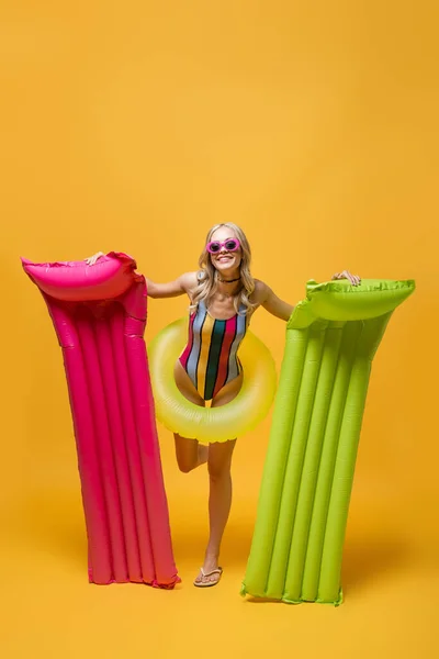 Full length of happy woman in sunglasses and swimsuit holding inflatable mattresses and standing with swim ring on yellow — Stock Photo