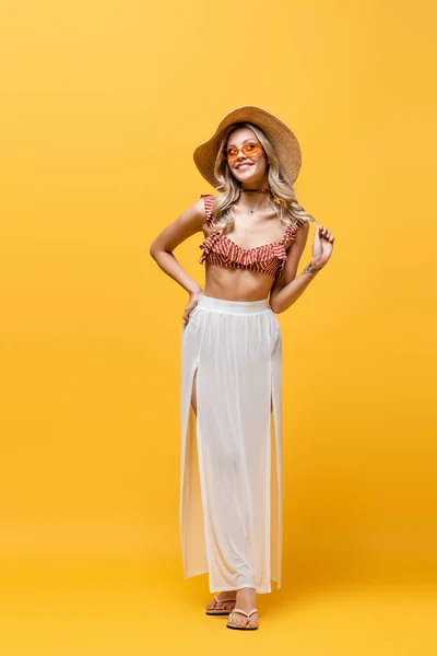 Full length of smiling woman in ruffle bikini top and white skirt standing with hand on hip on yellow — Stock Photo