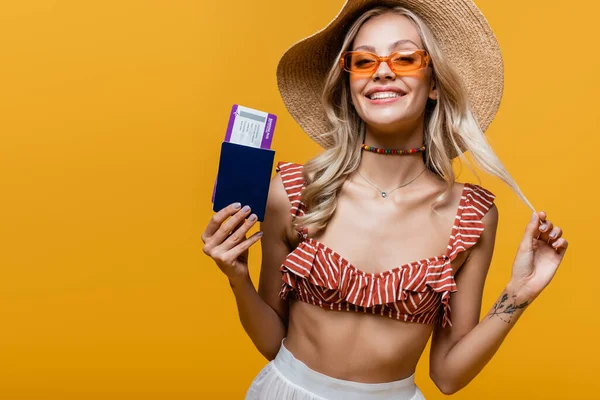 Tattooed woman in ruffle bikini top and sun hat smiling while holding passport with air ticket isolated on yellow — Stock Photo