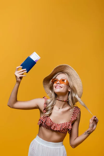 Cheerful woman in ruffle bikini top and sun hat smiling while holding passport with air ticket isolated on yellow — Stock Photo