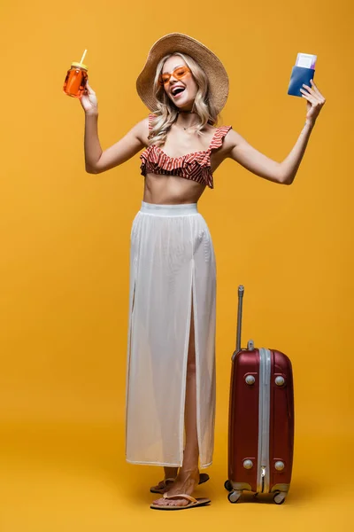 Полная длина счастливой женщины в шляпе, стоящей рядом с багажом, держа паспорт и коктейль на желтом — стоковое фото