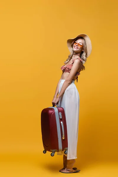 Full length of happy woman in sun hat lifting luggage on yellow — Stock Photo