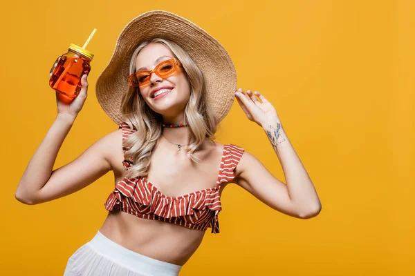 Lächelnde Frau mit Strohhut und Sonnenbrille mit Alkoholcocktail auf gelbem Hintergrund — Stockfoto