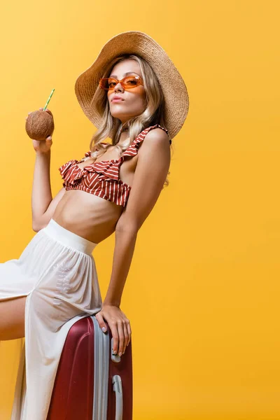 Mujer rubia en sombrero de sol sentada en el equipaje y sosteniendo coco con paja aislada en amarillo - foto de stock