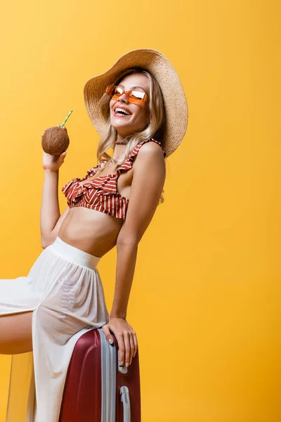 Весела жінка в сонячному капелюсі сидить на багажі і тримає кокос з соломою ізольовано на жовтому — стокове фото