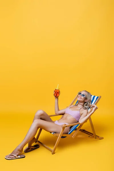 Happy young woman in swimsuit sitting on deck chair with cocktail on yellow — Stock Photo