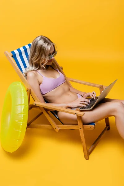 Femme blonde en lunettes de soleil et maillot de bain dactylographier sur ordinateur portable tout en étant assis dans la chaise longue sur jaune — Photo de stock