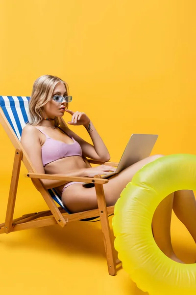 Jeune femme blonde en lunettes de soleil et maillot de bain en utilisant un ordinateur portable tout en étant assis sur une chaise longue sur jaune — Photo de stock
