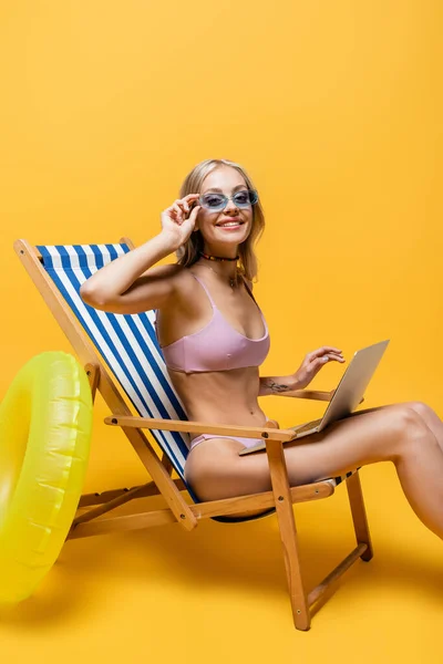 Femme souriante en lunettes de soleil et maillot de bain assis dans une chaise longue avec ordinateur portable sur jaune — Photo de stock