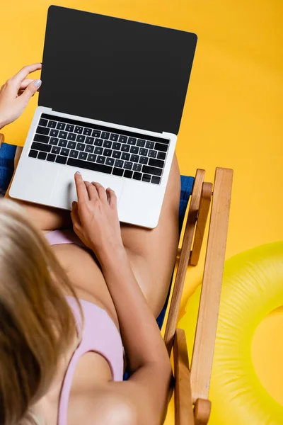 Vue du dessus de la femme blonde en maillot de bain en utilisant un ordinateur portable tout en étant assis sur une chaise longue sur jaune — Photo de stock
