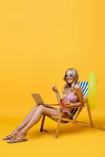 Blonde woman in sunglasses and swimsuit holding cocktail while sitting in deck chair with laptop on yellow — Stock Photo