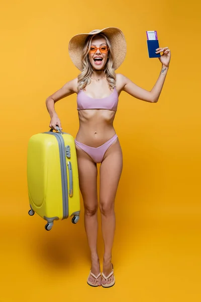 Volle Länge der staunenden Frau in Sonnenhut und Badeanzug, die mit Gepäck steht, während sie den Pass auf gelb hält — Stockfoto