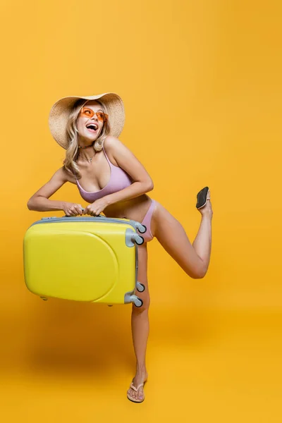 Full length of excited woman in straw hat and swimsuit standing with luggage on yellow — Stock Photo